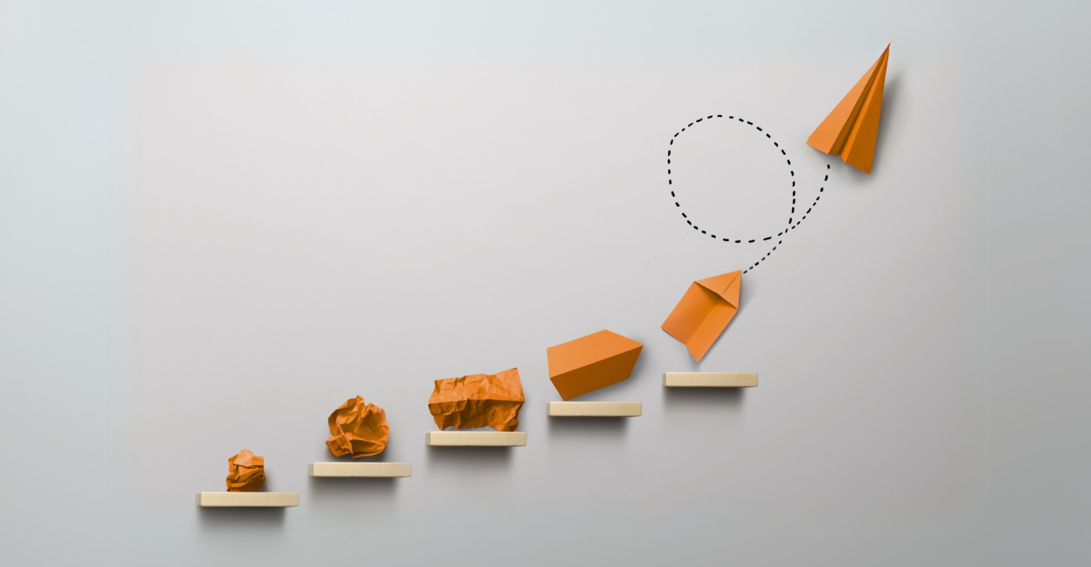 Mehrere Regalbretter mit zerknülltem Papier, gefaltetem Papier und Papierflugzeug; gestrichelte Fluglinie nach oben rechts