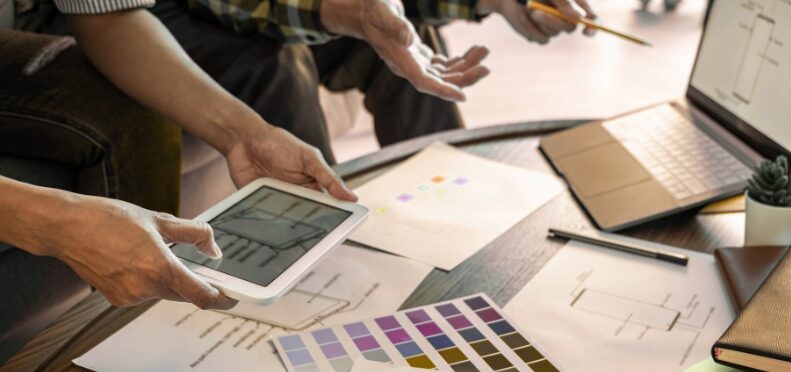 Mehrere Personen an Tisch mit Tablet, Laptop und Papieren; Farbmusterkarten und Diagramme liegen aus