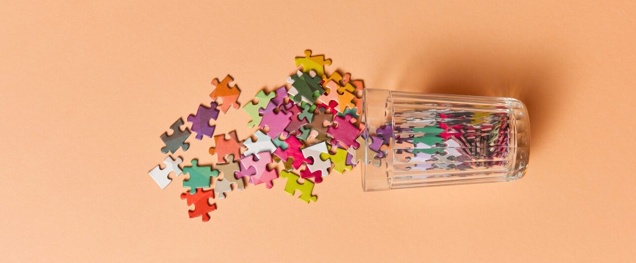 Umgestürztes Glas, aus dem bunte Puzzleteile auf eine einfarbige, orangefarbene Fläche herausfallen.