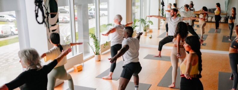 Mehrere Personen auf Yogamatten in Raum, Arme seitlich ausgestreckt, große Fenster an der Seite