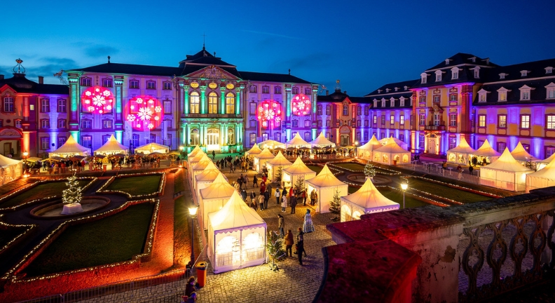 Beleuchteter Platz mit Gebäuden, Zelten und Menschen; bunte Lichtprojektionen an Fassaden bei Nacht