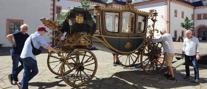 Vier Personen lehnen an einer reich verzierten Kutsche mit großen Rädern vor einem Schlossgebäude auf gepflastertem Platz.