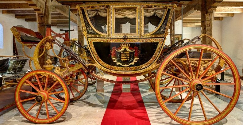 Verzierte historische Kutsche mit großen roten Rädern steht in einem Raum mit Holzbalken und rotem Teppich.