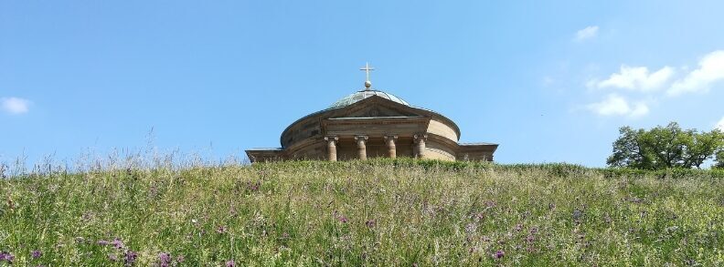 Runder Bau mit Säulen und Kuppel auf Hügel, davor Wiese mit Blumen, darüber blauer Himmel mit wenigen Wolken.