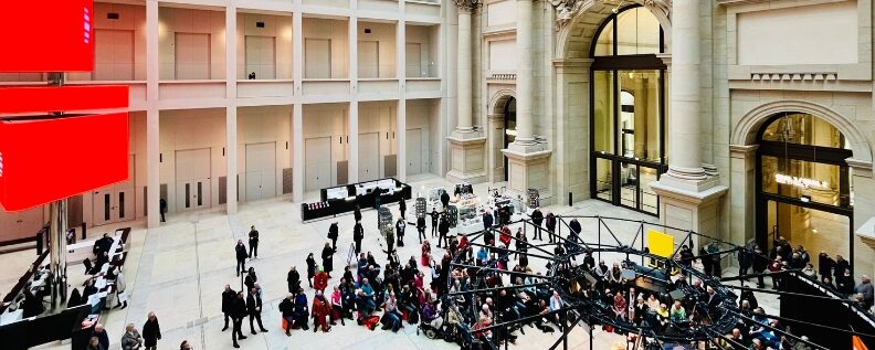 Großer Innenraum mit hoher Glasdecke, Säulen und Bögen; zahlreiche Personen stehen und sitzen um eine Bühne mit Technik.