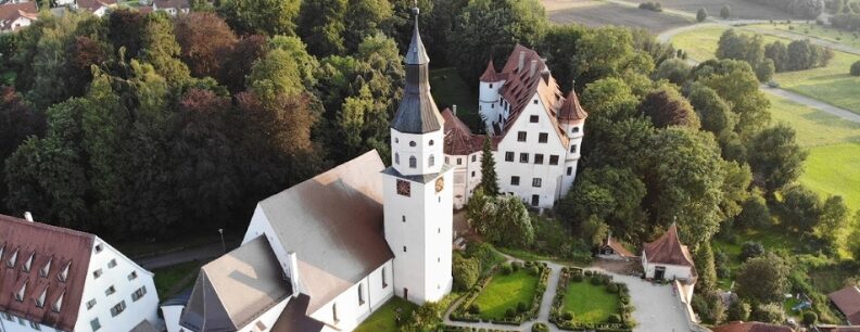 Luftaufnahme eines Gebäudekomplexes mit Kirche, Turm, Gartenanlagen und umliegenden Bäumen sowie weiteren Häusern.