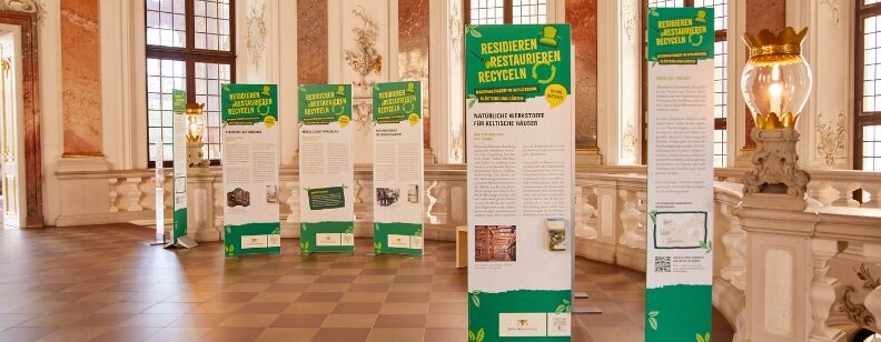 Mehrere Ausstellungstafeln mit Text und Überschrift „RESIDIEREN RESTAURIEREN RECYCELN“ stehen in einem Saal mit Fenstern und Säulen.