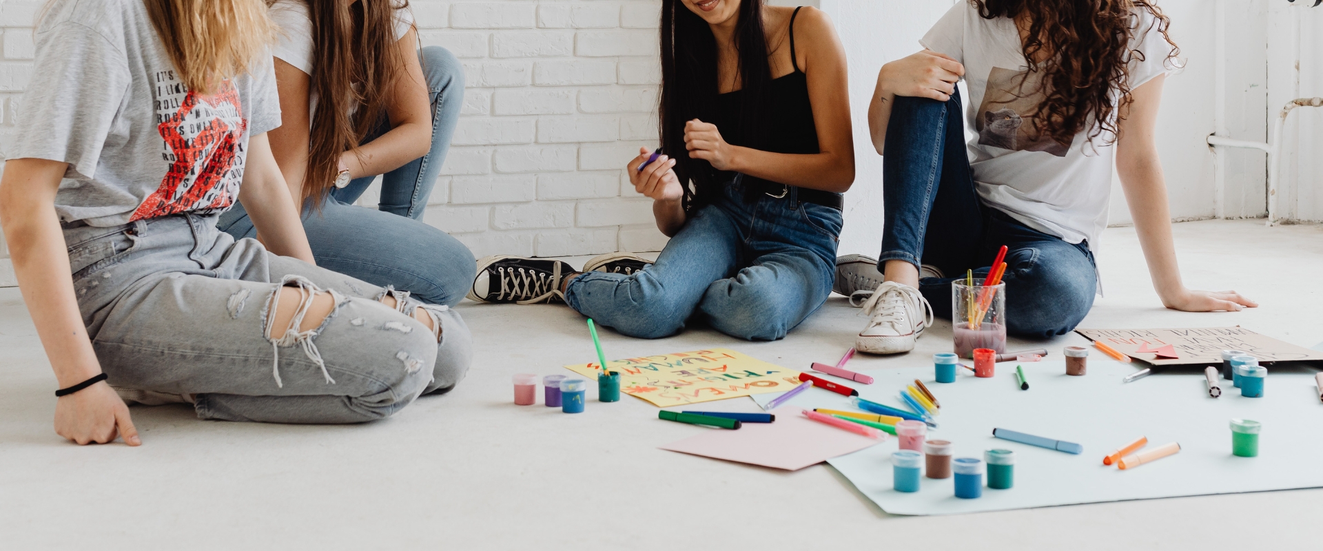 Mehrere Personen sitzen auf Boden mit Stiften und Farben; vor ihnen Papier mit Zeichnungen und Farbtöpfe