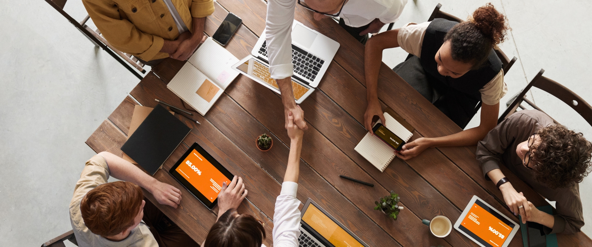 Mehrere Personen an Tisch mit Laptops und Tablets; zwei Hände in der Mitte beim Händedruck
