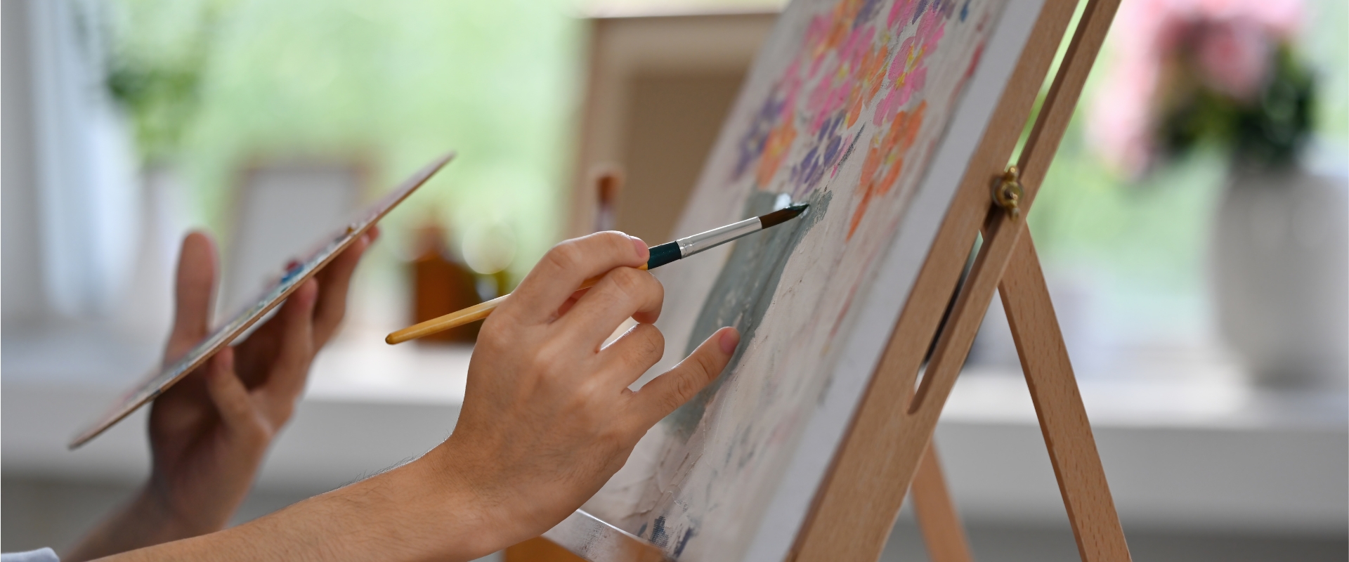Hand hält Pinsel und malt auf Leinwand auf Staffelei; zweite Hand hält Farbpalette