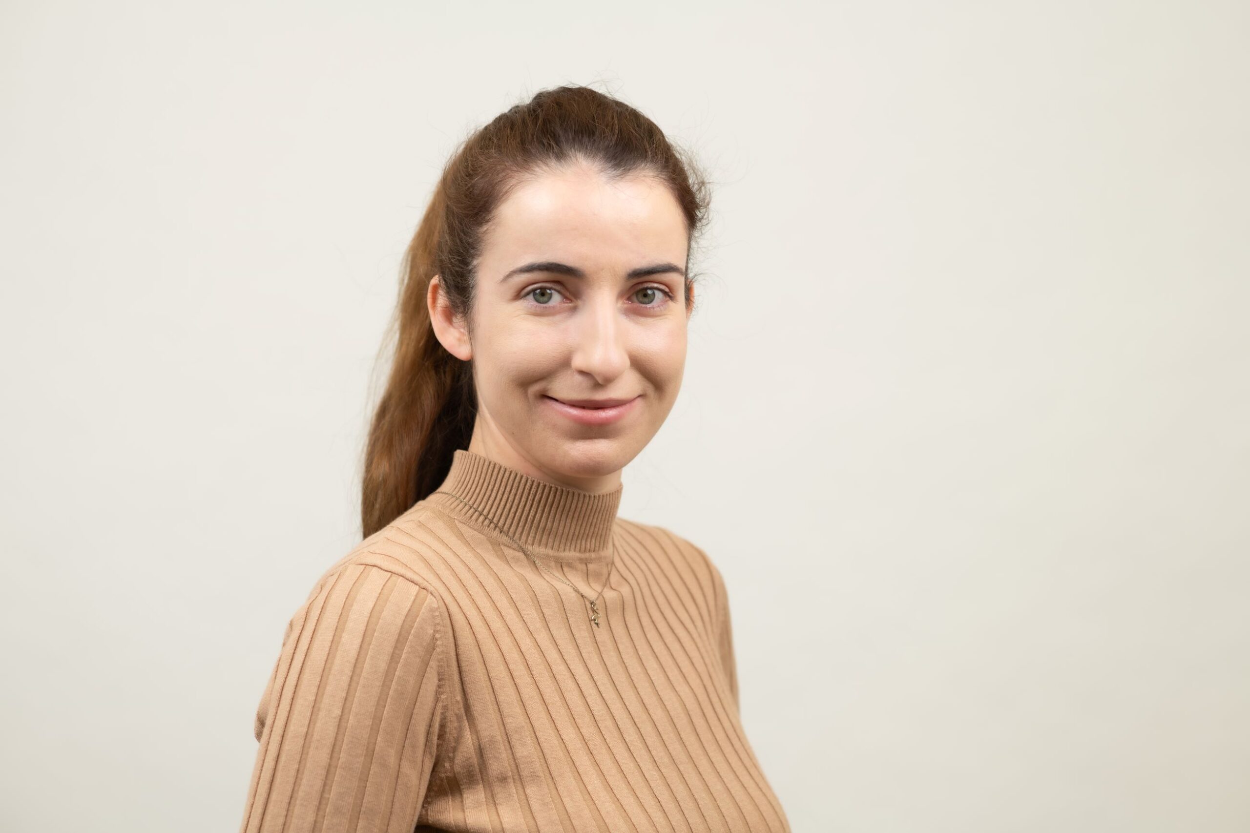 Eine Person mit zurückgebundenen Haaren trägt einen beigen Rollkragenpullover und blickt vor hellem Hintergrund in die Kamera.