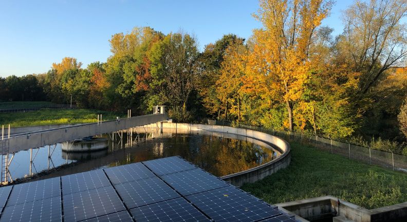 Rundes Wasserbecken mit Steg, Solarmodule im Vordergrund und Bäume mit Herbstlaub im Hintergrund