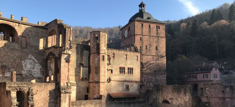 Burgruine mit Turm und Mauerwerk vor bewaldetem Hang unter blauem Himmel.