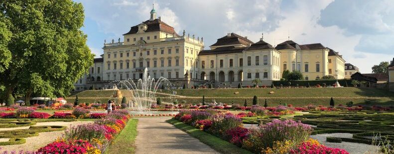 Schloss mit Springbrunnen und farbenreichen Blumenbeeten in barockem Garten unter bewölktem Himmel
